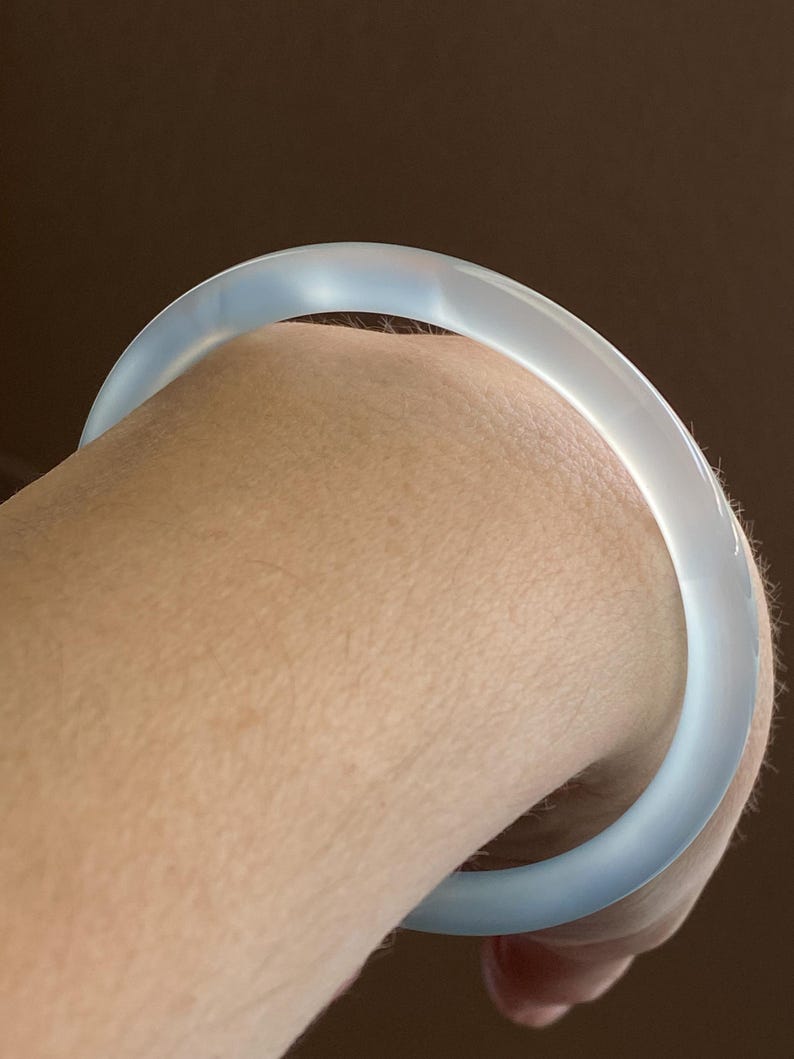 May include: A smooth, translucent, pale blue bangle bracelet with a rounded profile. The material appears semi-transparent, allowing light to pass through the surface, creating a soft glow and a consistent, polished finish around the entire circular band.