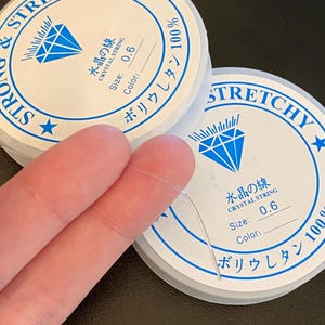 Op de afbeelding: Twee spoelen van helder, rekbaar kristallen draad, maat 0,6, met Japanse tekst en een diamantafbeelding. De spoelen zijn wit met blauwe tekst en een blauw diamantlogo. De tekst luidt "Strong & Stretchy" en "Crystal String".