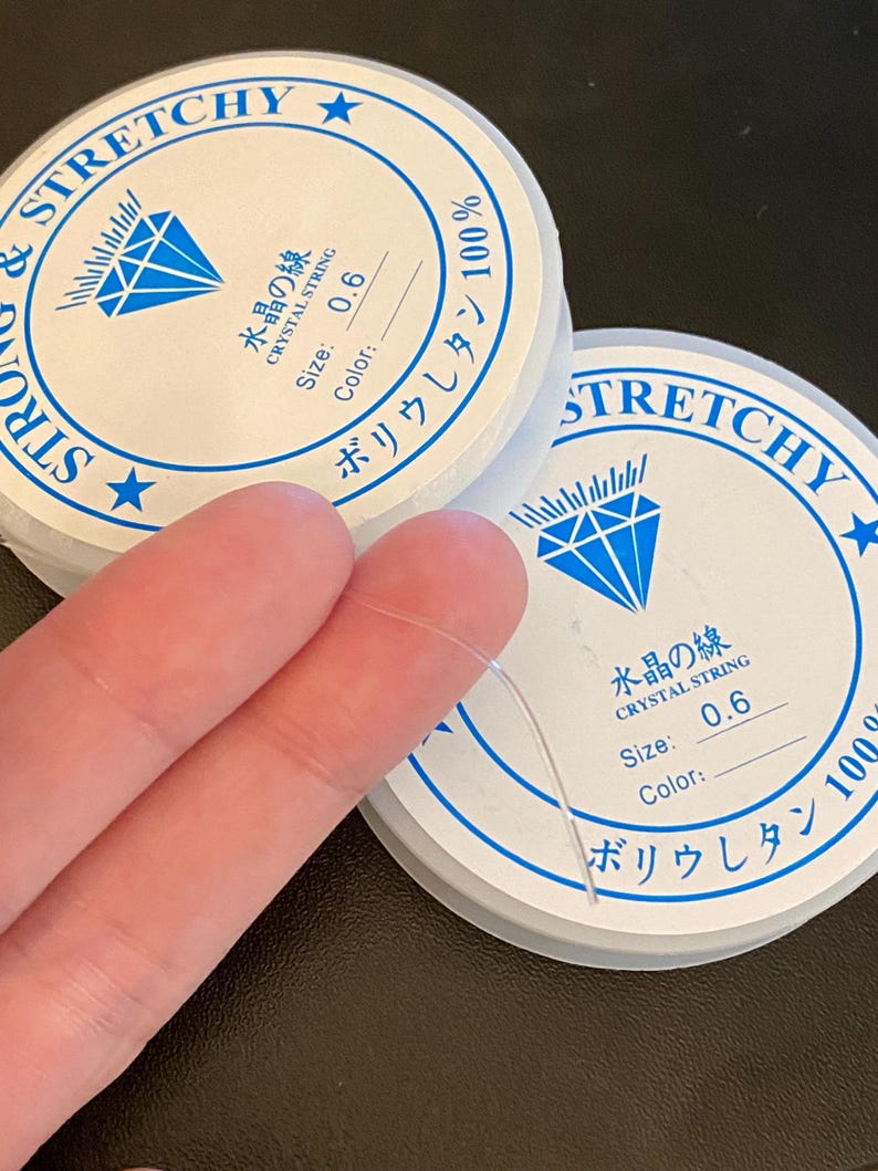 Op de afbeelding: Twee spoelen van helder, rekbaar kristallen draad, maat 0,6, met Japanse tekst en een diamantafbeelding. De spoelen zijn wit met blauwe tekst en een blauw diamantlogo. De tekst luidt "Strong & Stretchy" en "Crystal String".
