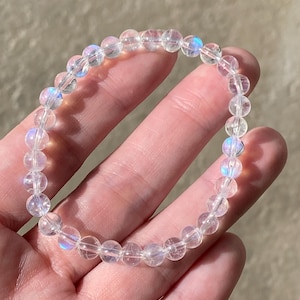 May include: A circular beaded bracelet composed of translucent, spherical beads. Each bead exhibits a clear, glass-like appearance with subtle internal light reflections that create a soft, iridescent blue glow when hit by direct light.
