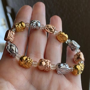 May include: A beaded bracelet featuring alternating gold, silver, and rose gold-colored charms. The charms have intricate designs and are strung on a clear, elastic band. The bracelet is a mix of metallic colors.