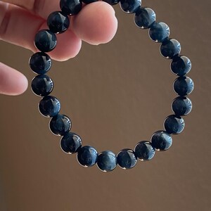 May include: A circular stretch bracelet composed of twenty-one polished, spherical beads. The beads feature a deep, dark blue colour with subtle, reflective chatoyant bands that create a shimmering effect across their smooth, rounded surfaces.