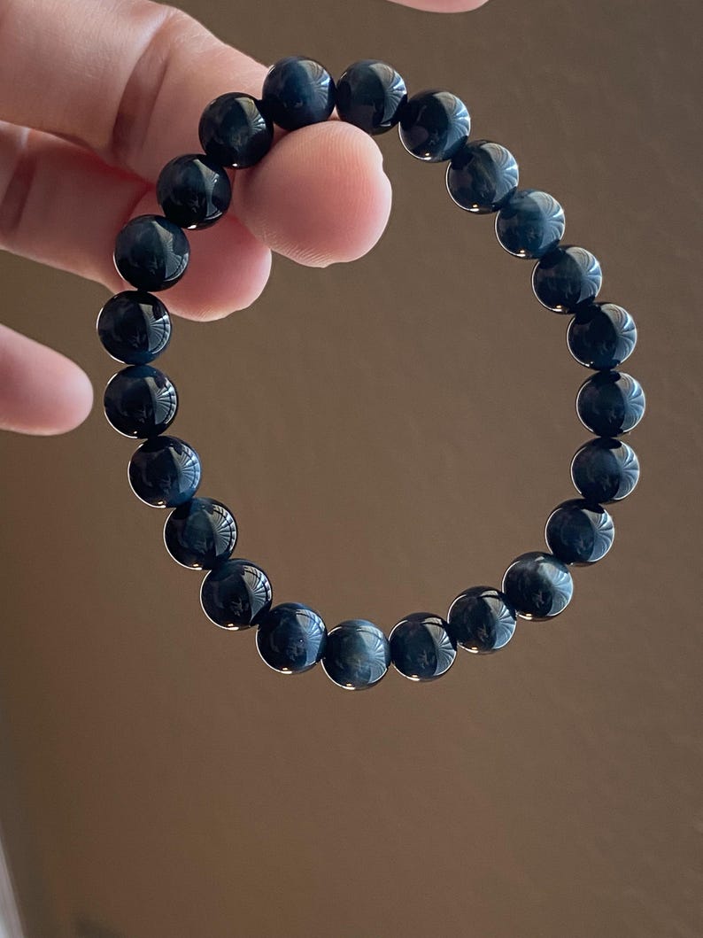 May include: A circular stretch bracelet composed of twenty-one polished, spherical beads. The beads feature a deep, dark blue colour with subtle, reflective chatoyant bands that create a shimmering effect across their smooth, rounded surfaces.