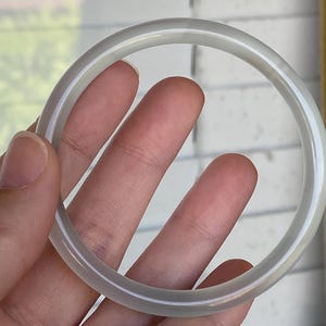 May include: A smooth, circular bangle bracelet crafted from a translucent, pale grey stone material. The bangle features a polished, rounded surface with a consistent thickness throughout its entire circumference, showcasing a subtle, cloudy appearance.