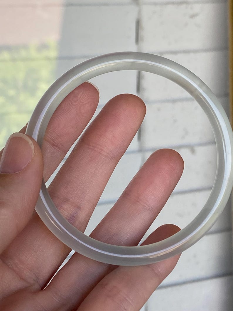 May include: A smooth, circular bangle bracelet crafted from a translucent, pale grey stone material. The bangle features a polished, rounded surface with a consistent thickness throughout its entire circumference, showcasing a subtle, cloudy appearance.