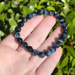 May include: A stretch bracelet composed of round, polished dark blue tiger eye beads. Each bead displays a distinct chatoyant light reflection, creating a shimmering, cat-eye effect across the surface of the deep blue stone spheres.