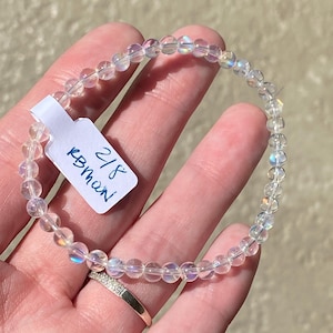 May include: A clear, iridescent beaded bracelet. The bracelet is made of small, round beads strung together. A small white tag with the text "2/8 KBman" is attached to the bracelet. The bracelet is worn on a wrist.