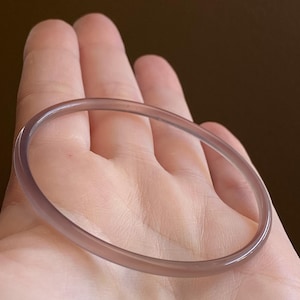 May include: A translucent, lavender-colored bangle bracelet. The smooth, circular bracelet is held in a person's hand, showcasing its simple, elegant design. The bracelet's color is a soft, muted purple, and it appears to be made of a polished stone or glass.