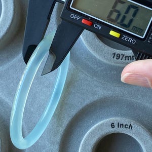 May include: A smooth, translucent light blue bangle bracelet being measured by a black digital caliper. The digital display on the caliper clearly shows a measurement of 6.0 mm. The bangle has a rounded, polished surface and a consistent thickness.