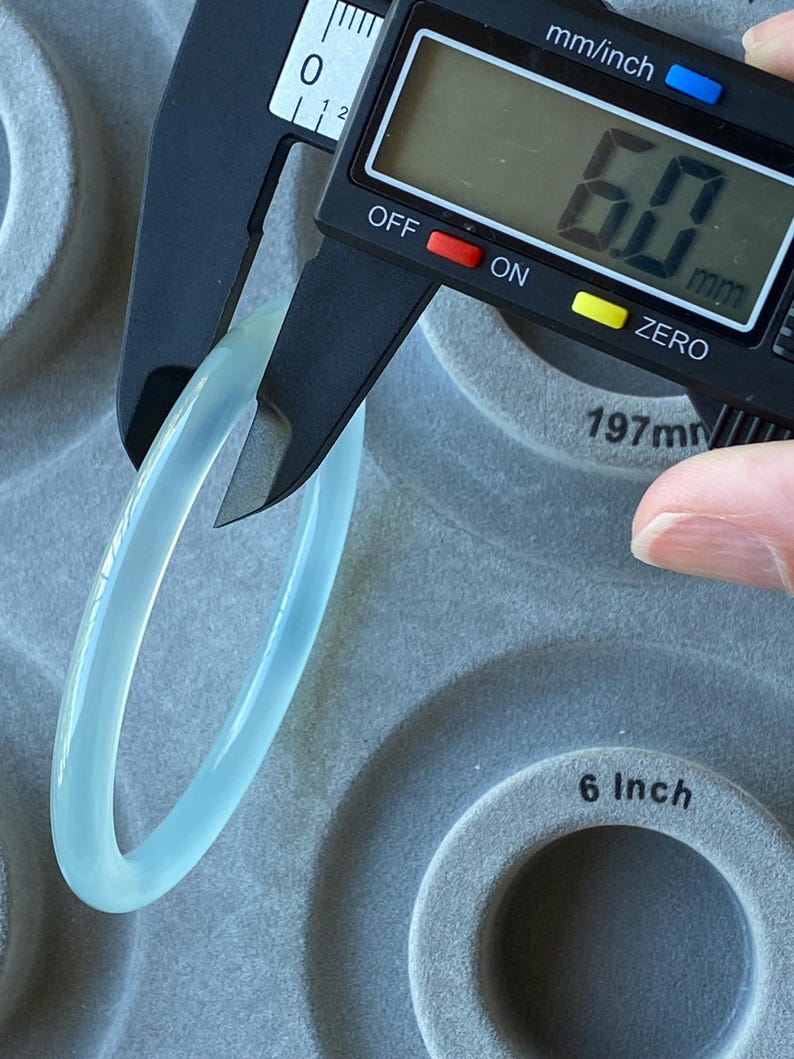 May include: A smooth, translucent light blue bangle bracelet being measured by a black digital caliper. The digital display on the caliper clearly shows a measurement of 6.0 mm. The bangle has a rounded, polished surface and a consistent thickness.