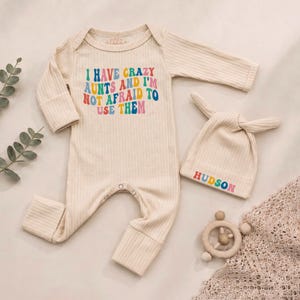 May include: A cream-colored ribbed long-sleeve romper and matching knotted hat. The romper features the colorful text I HAVE CRAZY AUNTS AND I'M NOT AFRAID TO USE THEM. The hat displays the name HUDSON in matching colorful, retro-style lettering.