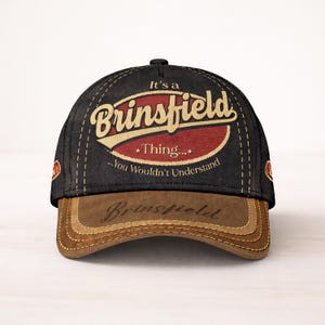 May include: Black and brown baseball cap with the name "Brinsfield" in a vintage style. The cap features the text "It's a Brinsfield Thing... You Wouldn't Understand" in a retro design.