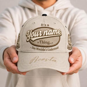Op de afbeelding: Een beige honkbalpet met de tekst It's a your name Thing... You Wouldn't Understand in bruine letters binnen een ovaal kader. De klep toont de naam Acosta in cursief schrift en de zijpanelen zijn voorzien van cirkelvormige emblemen.