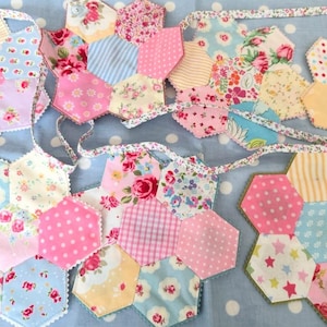Peut inclure: Guirlande de fanions en tissu faite à la main avec un motif patchwork. Elle est composée de divers tissus à motifs floraux et autres, dans des tons de rose, bleu, jaune et blanc. Les pièces hexagonales sont cousues avec un ruban décoratif.