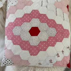 May include: Square patchwork pillow with a geometric design. The pillow features a central red hexagon surrounded by white, pink polka dot, and patterned fabric hexagons. The pillow is resting on a fluffy white surface.
