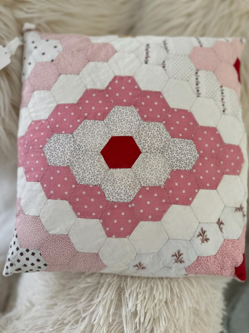 Op de afbeelding: Vierkant patchwork kussen met een geometrisch ontwerp. Het kussen heeft een centrale rode zeshoek omgeven door witte, roze stippen en patroonstof zeshoeken. Het kussen ligt op een pluizig wit oppervlak.