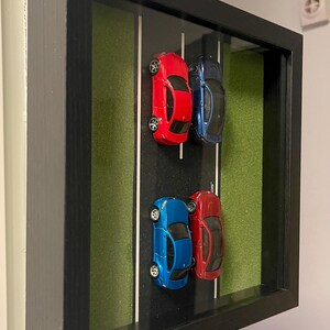 May include: A black framed display with four toy cars, two red and two blue, arranged on a black surface with white lines, simulating a race track. The background is green. A decorative item.