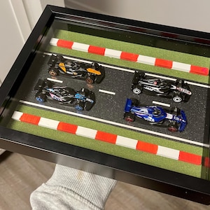 Puede incluir: Una exhibición enmarcada con cuatro coches de carreras en miniatura en una pista. Los coches son negros, azules y naranjas, con logotipos de patrocinadores. La pista incluye césped verde, líneas blancas y detalles de bordillos rojos y blancos. El marco es negro.