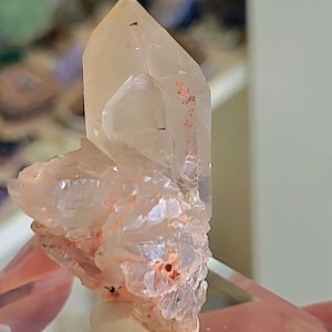 May include: A clear quartz crystal cluster with a large, pointed main crystal and smaller, attached crystals. The quartz has a pale, translucent color with reddish-orange inclusions and a rough, textured base. The crystal is set against a blurred background.