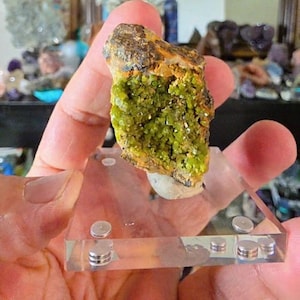 May include: A vibrant green mineral specimen with a rough, textured surface, mounted on a clear acrylic display stand. The mineral has a mix of green and yellow hues, with a dark brown top.