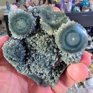 May include: A close-up of a hand holding a cluster of green quartz crystals. The quartz has a bubbly texture with circular patterns and a dark green color. The crystal formation is a natural, organic shape.