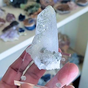 May include: A clear quartz crystal with a pointed tip, held in a clear acrylic stand. The crystal has a rough, textured surface with small, sparkling inclusions and some green and gray mineral deposits. Other crystals are visible in the background.