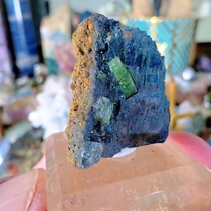 May include: A raw mineral specimen featuring a dark blue, crystalline structure with embedded bright green, translucent crystals. One side of the stone has a textured, tan-colored matrix with small, rounded, brownish-orange mineral deposits.