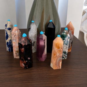 May include: A collection of nine hexagonal crystal towers in various colors and patterns, including blue and white, peach, black, clear, purple, and banded orange, each topped with a small, bright turquoise blue dot of adhesive material.