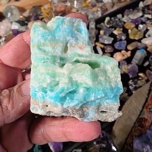 May include: A raw, unpolished mineral specimen featuring horizontal bands of vibrant turquoise, pale mint green, and white. The stone has a rough, textured surface with natural crevices and a solid, earthy base layer of gray and tan mineral deposits.
