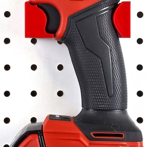 May include: A red and black Milwaukee FUEL impact driver with a black battery pack. The tool is mounted on a white pegboard with red brackets. The Milwaukee logo is prominently displayed.