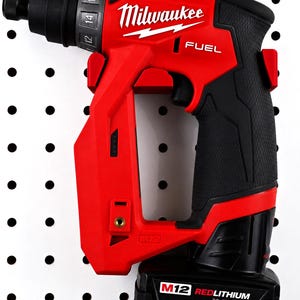 May include: A red and black Milwaukee FUEL M12 cordless screwdriver. The tool has a black handle and a battery pack labeled "M12 REDLITHIUM XC6.0". The Milwaukee logo is visible in white. The screwdriver is mounted on a white pegboard.