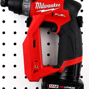 May include: A red and black Milwaukee FUEL M12 cordless screwdriver. The tool is mounted on a white pegboard. The screwdriver has a black handle and a battery pack labeled "M12 REDLITHIUM XC6.0". The Milwaukee logo is visible.