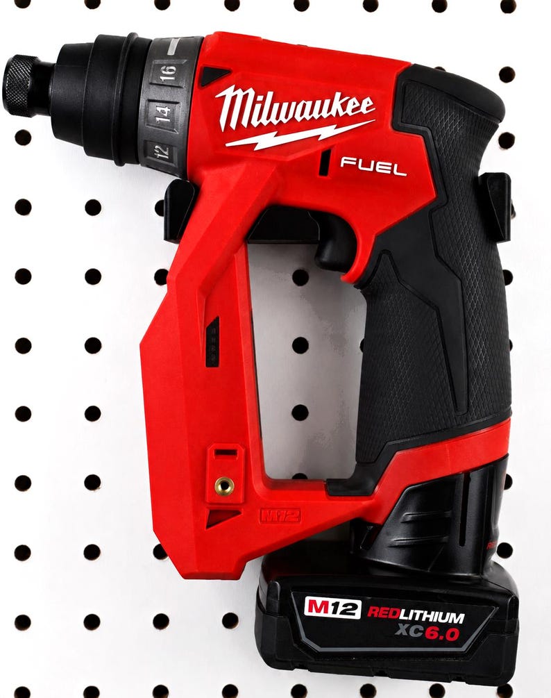 May include: A red and black Milwaukee FUEL M12 cordless screwdriver. The tool is mounted on a white pegboard. The screwdriver has a black handle and a battery pack labeled "M12 REDLITHIUM XC6.0". The Milwaukee logo is visible.