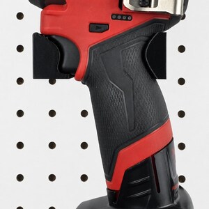 May include: A red and black Milwaukee FUEL impact driver. The tool has a black grip, a red body, and the Milwaukee logo in white. The battery pack is labeled M12 REDLITHIUM XC 6.0. It is mounted on a black pegboard.