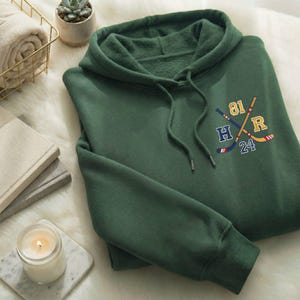 May include: A forest green hoodie with a drawstring hood and a kangaroo pocket. The hoodie features an embroidered design of crossed hockey sticks with the numbers 81 and 24, and the letters H and R. The hoodie is made of a soft, comfortable material.