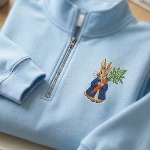 May include: A light blue sweatshirt with a quarter-zip neckline. The sweatshirt features an embroidered image of a rabbit wearing a blue coat and holding a carrot and greenery. The sleeves and neckline are ribbed.