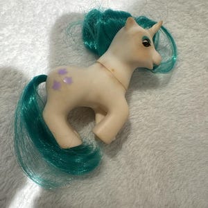 Puede incluir: Un juguete de poni vintage con cuerpo color crema, melena, cola y detalles de ojos turquesa. El poni tiene un solo cuerno y marcas moradas en forma de flor en el flanco. El juguete está sobre una superficie blanca texturizada.