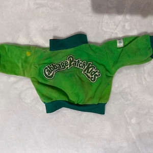 Pode incluir: Uma camisola de manga comprida verde brilhante com acabamentos em verde escuro na gola, punhos e bainha. As costas apresentam o texto Cabbage Patch Kids bordado numa fonte estilizada e uma pequena etiqueta branca na manga esquerda.