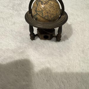 Puede incluir: Un sacapuntas de globo vintage. El globo es de color beige con detalles geográficos azules y verdes. Está montado sobre un soporte de metal oscuro con cuatro patas y una base de sacapuntas. El globo se sujeta con un anillo de metal.