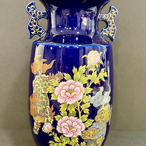 May include: A cobalt blue ceramic vase with a glossy finish. The vase features pink peony flowers, green leaves, and mythical creatures in gold, orange, and gray. The vase has two handles and measures approximately 12 inches tall.