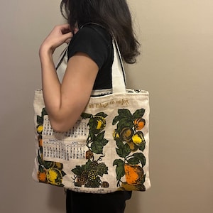 May include: A beige tote bag with a fruit and calendar design. The bag features images of oranges, lemons, and grapes, with a calendar grid and the text "all things to enjoy". The bag has white straps.
