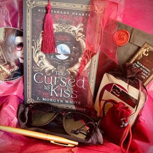 May include: A book titled Too Cursed to Kiss by J. Morgyn White with a red tassel bookmark, black sunglasses, a gold pen, a red velvet pouch, a wax-sealed envelope, and a sticker featuring a person with glasses and the text Time is precious.
