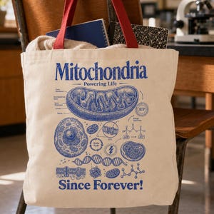 Puede incluir: Un bolso de tela beige con asas rojas que presenta un estampado gráfico azul de mitocondrias, estructuras celulares y el texto Mitochondria Powering Life Since Forever, junto con diagramas biológicos detallados y fórmulas.