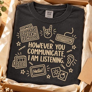Könnte beinhalten: Ein dunkelgraues T-Shirt mit einem hellbeigen Grafikdruck und dem Text "HOWEVER YOU COMMUNICATE, I AM LISTENING." Das Design zeigt Symbole für ein Kommunikationsboard, eine Handgeste, eine Computertastatur, ein Ohr und ein Tablet.