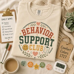 Op de afbeelding: Een crèmekleurig T-shirt met korte mouwen en een grafische print met de tekst Behavior Support Club in rode, groene en gele letters. Het ontwerp bevat een smiley, een icoon van een klembord, hartjes en bladeren op de voorzijde van het shirt.