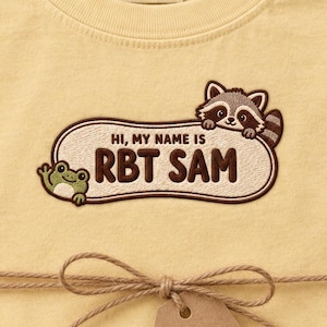 Könnte beinhalten: Ein blassgelbes T-Shirt mit einem gestickten Aufnäher, der einen Waschbären und einen Frosch zeigt. Der Aufnäher enthält den Text "HI, MY NAME IS RBT SAM" in brauner Schrift. Das T-Shirt hat ein Comfort Colors 1717 Etikett in Größe L und ist mit Bindfaden verschnürt.