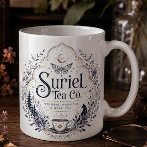 Könnte beinhalten: Ein weißer Keramikbecher mit einer dekorativen Illustration und dem Text Suriel Tea Co. Whispers, Warnings & Warm Tea. Das Design zeigt einen Nachtfalter, eine Mondsichel, eine kleine Teetasse, eine Berglandschaft und einen Fuchs, umrahmt von Ranken.