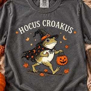 May include: A charcoal gray short-sleeve t-shirt featuring a graphic of a frog wearing a witch hat and cape, carrying a jack-o'-lantern. The text HOCUS CROAKUS is printed above the frog, surrounded by orange stars, crescent moons, and autumn leaves.