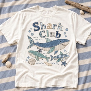 Puede incluir: Una camiseta blanca de manga corta con un gráfico de tres tiburones y el texto Shark Club. El diseño utiliza varios tejidos estampados en azul y blanco con costuras visibles, acompañado de pequeñas ilustraciones de conchas y una estrella de mar.