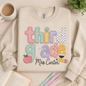 May include: A beige crewneck sweatshirt featuring the text "third grade" in colorful, patterned, stitched-style letters. A pink apple graphic is integrated into the letter "g," and a yellow pencil graphic is positioned below the text "Mrs. Carter."